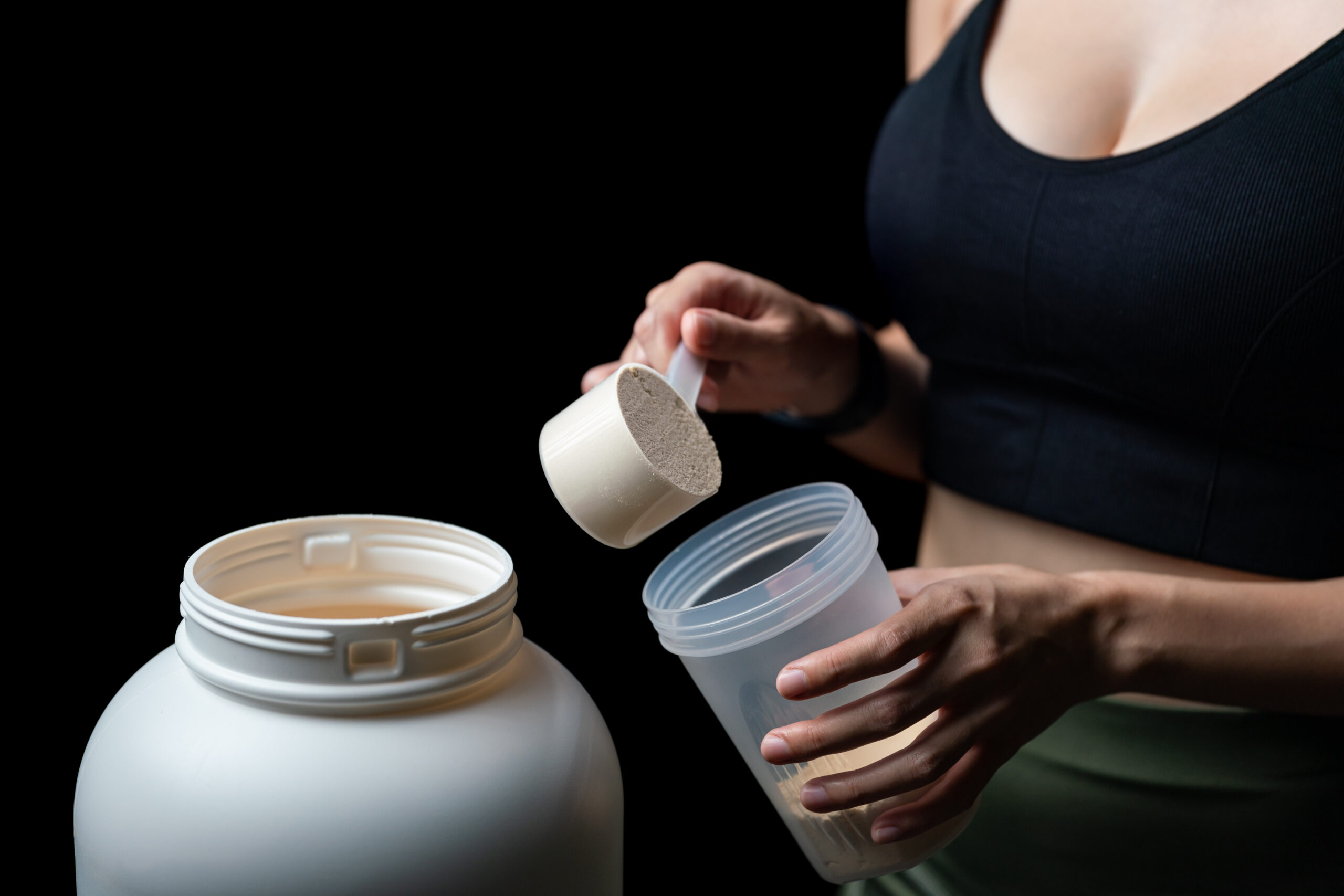 Close up of women with measuring scoop of whey protein, jar and shaker bottle, preparing protein shake.